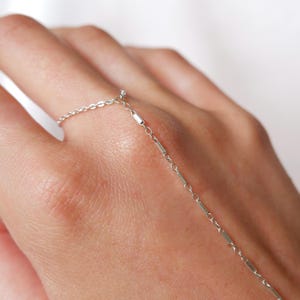 May include: A delicate silver ring connected by a chain to a bracelet. The ring is on a finger, and the chain drapes across the back of the hand. The jewelry has a minimalist design, with small rectangular and circular links.
