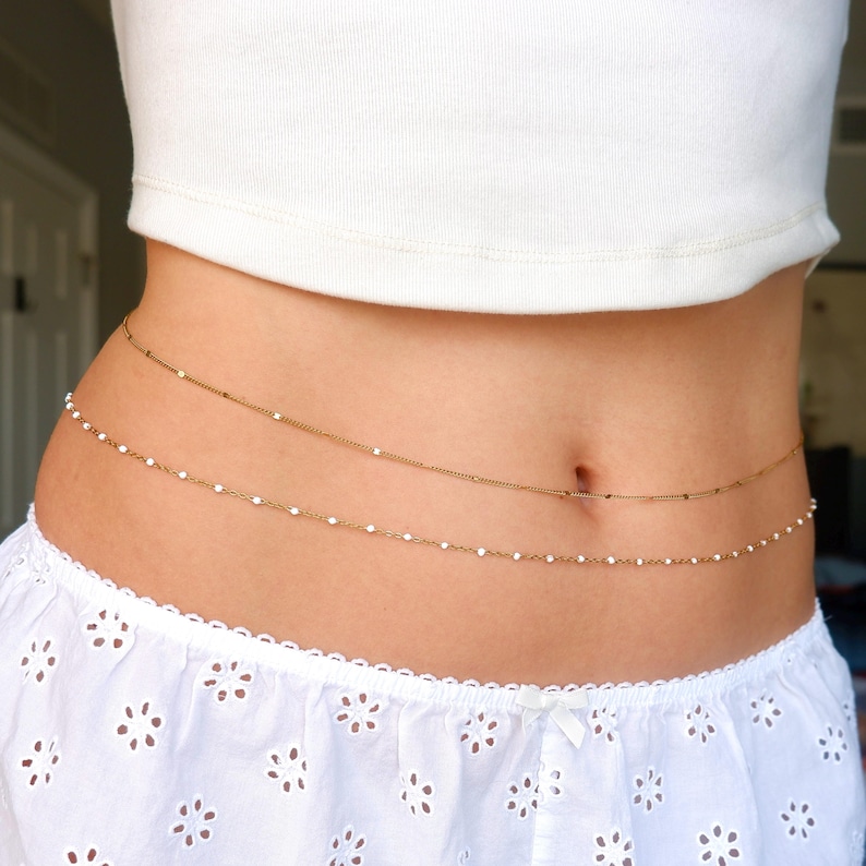 Peut inclure: Une cha&icirc;ne de ventre dor&eacute;e &agrave; double rang&eacute;e compos&eacute;e d'une cha&icirc;ne fine simple et d'une cha&icirc;ne orn&eacute;e de petites perles blanches r&eacute;guli&egrave;rement espac&eacute;es. Le bijou repose sur la taille, entre un haut blanc et un short blanc.