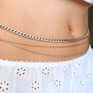 Peut inclure: Un ensemble de trois cha&icirc;nes de taille superpos&eacute;es comprenant une cha&icirc;ne gourmette argent&eacute;e plus &eacute;paisse et deux cha&icirc;nes plus fines et d&eacute;licates, l'une dor&eacute;e et l'autre argent&eacute;e. Elles sont port&eacute;es sur un tissu blanc &agrave; motif floral ajour&eacute;.