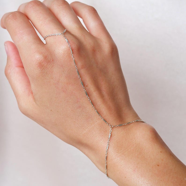 May include: A silver hand chain bracelet. The delicate jewelry piece connects a ring to a bracelet with a fine chain. The chain is made of small, rectangular links. The bracelet is worn on a hand with a light background.