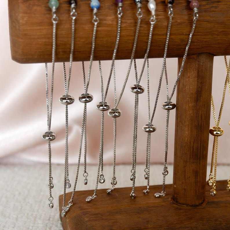 Puede incluir: Un expositor de pulseras ajustables de cadena plateada y dorada con peque&ntilde;os detalles de cristal. Las pulseras est&aacute;n colgadas de un soporte de madera.