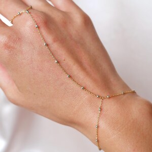 May include: A delicate gold hand chain with small silver beads. The chain connects a ring to a bracelet, draping across the back of the hand. The jewelry piece is designed with a fine chain and small, evenly spaced beads.