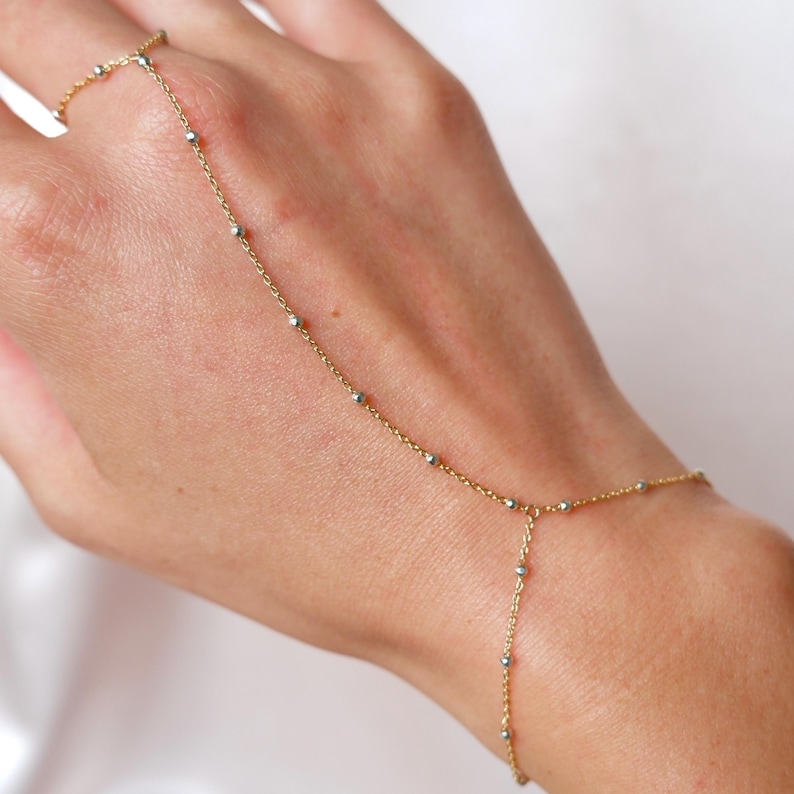 May include: A delicate gold hand chain with small silver beads. The chain connects a ring to a bracelet, draping across the back of the hand. The jewelry piece is designed with a fine chain and small, evenly spaced beads.
