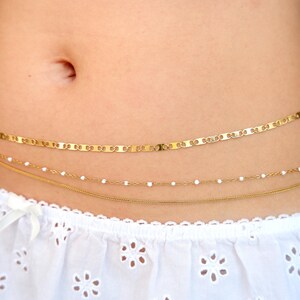 Peut inclure: Une cha&icirc;ne de taille dor&eacute;e &agrave; trois rangs compos&eacute;e d'une cha&icirc;ne &agrave; maillons plats, d'une cha&icirc;ne d&eacute;licate avec de petites perles blanches et d'une fine cha&icirc;ne serpent. Le bijou est port&eacute; sur un tissu blanc &agrave; motif floral ajour&eacute;.
