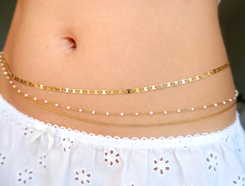 Peut inclure: Une cha&icirc;ne de taille dor&eacute;e &agrave; trois rangs compos&eacute;e d'une cha&icirc;ne &agrave; maillons plats, d'une cha&icirc;ne d&eacute;licate avec de petites perles blanches et d'une fine cha&icirc;ne serpent. Le bijou est port&eacute; sur un tissu blanc &agrave; motif floral ajour&eacute;.