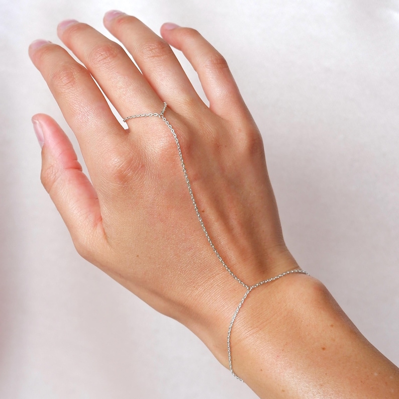 May include: A silver hand chain bracelet. The delicate jewelry piece connects a ring to a bracelet with a fine chain. The ring is on the index finger, and the bracelet is on the wrist. The chain is draped across the back of the hand.