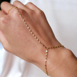 May include: A delicate gold hand chain featuring a ring, a bracelet, and a chain that connects them. The chain is adorned with small, daisy-like charms. The jewelry is worn on a hand against a white background.