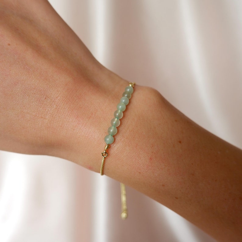 Puede incluir: Un delicado brazalete de cadena dorada con una fila de cuentas de color verde claro. El brazalete es ajustable y tiene un cierre deslizante.