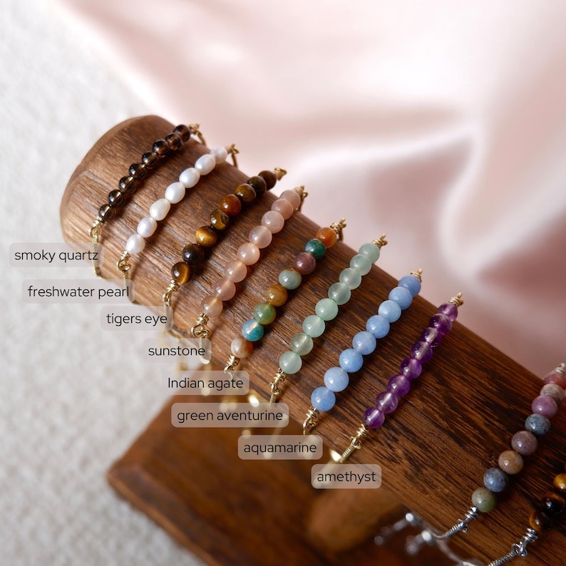 Puede incluir: Un conjunto de 10 pulseras de piedras preciosas con cierres dorados. Las pulseras est&aacute;n dispuestas en fila sobre una superficie de madera. Las piedras preciosas son cuarzo ahumado, perla de agua dulce, ojo de tigre, piedra de sol, &aacute;gata india, aventurina verde, aguamarina, amatista y m&aacute;s.