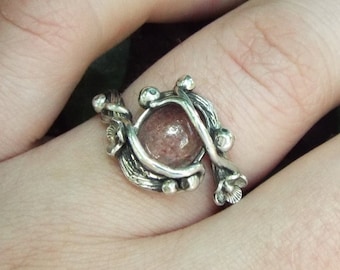 May include: A silver ring with a pink stone set in a delicate, twig-like design. The ring is worn on a person's finger.