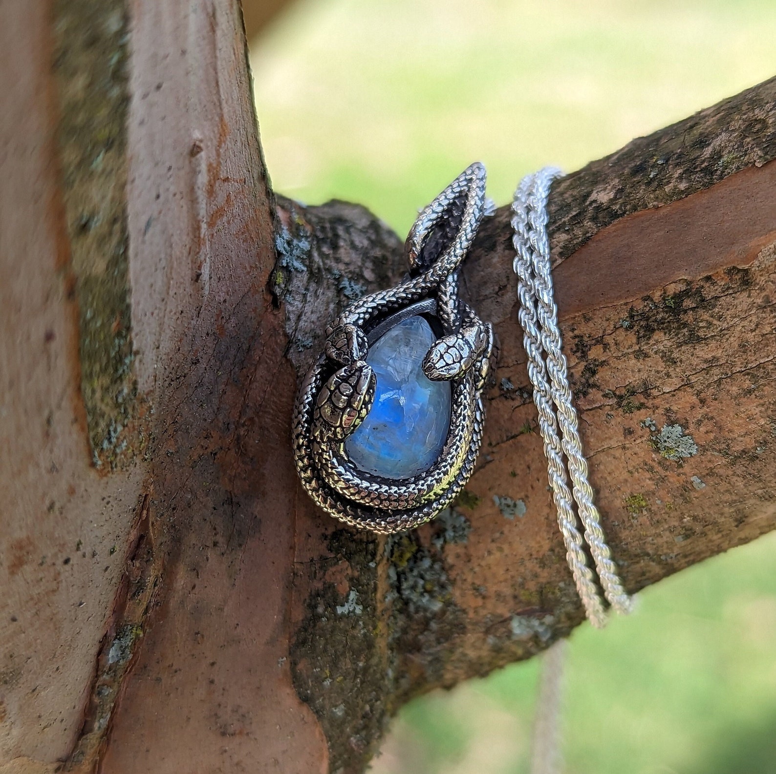 Sterling silver snake pendant with teardrop moonstone coiled around it, hanging on tree bark.