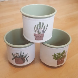 May include: Three white fabric-covered pots with green rims. Each pot has a different embroidered design of a potted plant with a small bird. The plants are labelled "Rosemary", "Thyme", and "Mint".