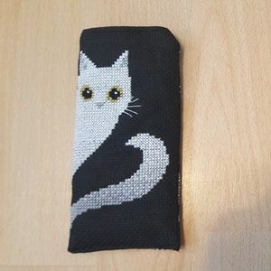 以下が含まれることがあります： 黒い布製の眼鏡ケースで、前面に白い猫が刺繍されています。猫は黄色い目をしており、白い尻尾をしています。