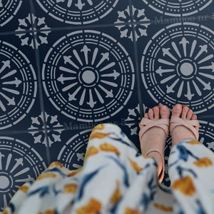 Op de afbeelding: Een sjabloon met een herhalend patroon van cirkelvormige ontwerpen met een centrale cirkel en stralende lijnen. De sjabloon bevindt zich op een donkerblauwe vloer en de voeten van een persoon zijn zichtbaar in het midden van de afbeelding. De tekst "Mamboo nr 5" is zichtbaar in de rechterbenedenhoek van de afbeelding.