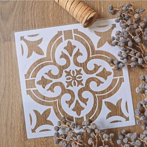 Pode incluir: Estêncil de plástico branco com um padrão de ladrilho decorativo. O estêncil está sobre uma superfície de madeira.