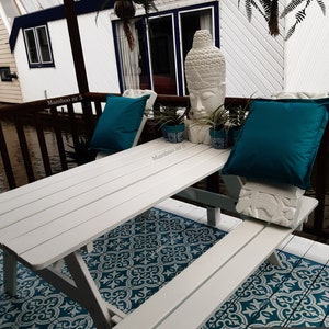 May include: White wooden picnic table with two benches, set on a teal and white patterned outdoor rug. A white Buddha statue sits on the table with two teal cushions and potted plants.