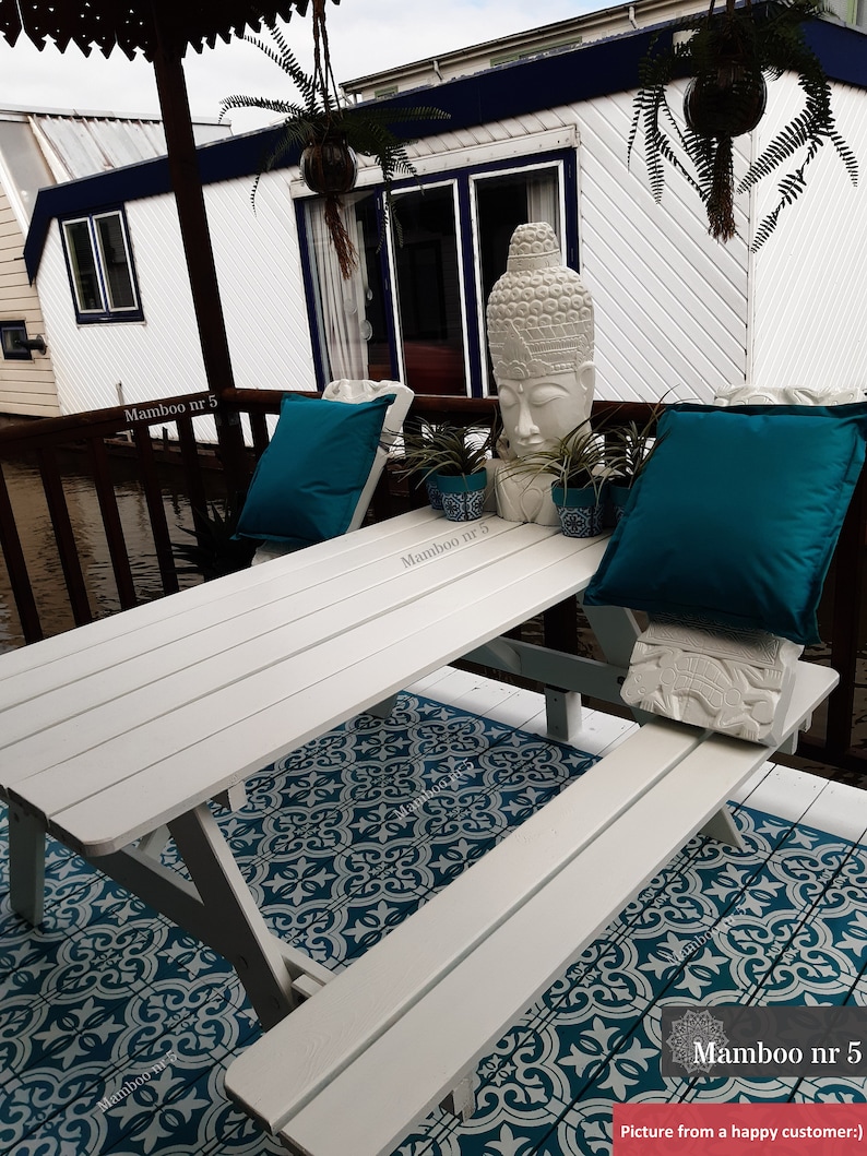 May include: White wooden picnic table with two benches, set on a teal and white patterned outdoor rug. A white Buddha statue sits on the table with two teal cushions and potted plants.