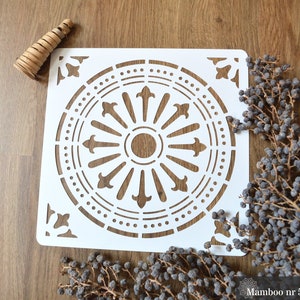 May include: White stencil with a decorative circular design. The design features a central circle with a smaller circle inside, surrounded by a ring of pointed shapes. The outer ring of the design is a circle of dots.