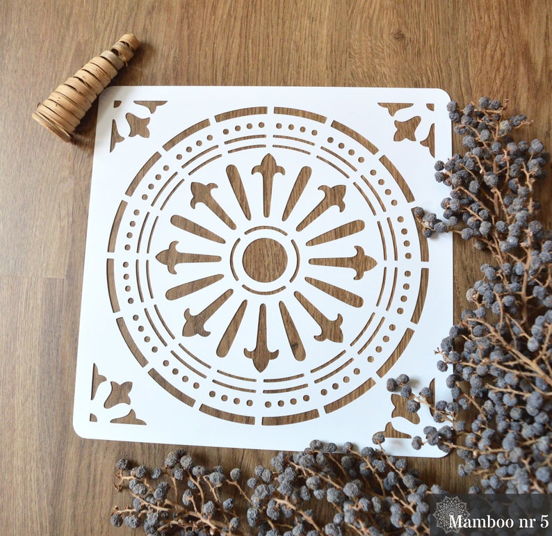 May include: White stencil with a decorative circular design. The design features a central circle with a smaller circle inside, surrounded by a ring of pointed shapes. The outer ring of the design is a circle of dots.