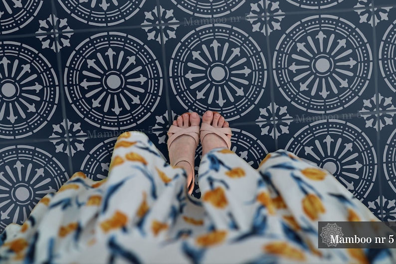 May include: A stencil with a repeating pattern of circular designs with a central circle and radiating lines. The stencil is on a dark blue floor and a person's feet are visible in the center of the image. The text "Mamboo nr 5" is visible in the bottom right corner of the image.