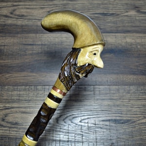 Pode incluir: Uma bengala de madeira com uma cabeça esculpida de um homem com barba e chapéu. A bengala é decorada com faixas marrons, brancas e de cobre.