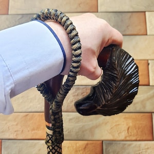 Può includere: Un bastone da passeggio in legno con un manico scolpito a forma di testa di animale. Il bastone è avvolto da un cordoncino intrecciato nero e marrone.