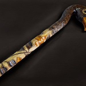 May include: A wooden walking stick with a carved duck head handle. The duck's head is brown with a yellow beak and black eyes. The stick is decorated with carved leaves and flowers.