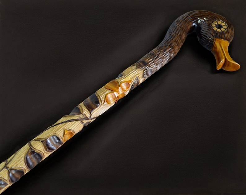 May include: A wooden walking stick with a carved duck head handle. The duck's head is brown with a yellow beak and black eyes. The stick is decorated with carved leaves and flowers.