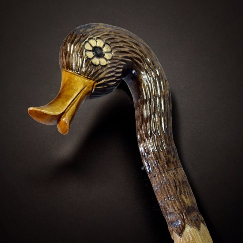 May include: A wooden walking stick carved in the shape of a duck's head. The duck's head is brown and black with a yellow beak and a white flower design on the top of the head.