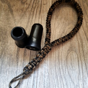 Könnte beinhalten: Eine schwarze und braune Paracord-Handschlaufe mit einer Schlaufe, neben zwei schwarzen Gummistöpseln für einen Gehstock. Das Paracord ist in einem Muster aus Schwarz und Beige gewebt. Die Stöpsel sind zylindrisch.