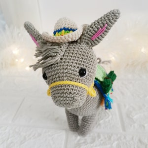 A handmade grey crochet donkey wearing a small white hat with a rainbow brim. The donkey has black eyes, pink inner ears, and a yellow bridle. A green and blue pack is on its back.