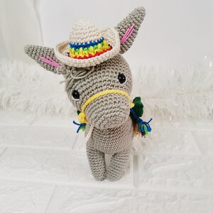 A crocheted grey donkey wearing a small sombrero with a rainbow-coloured brim. The donkey has a yellow bridle and colourful tassels. The ears have pink accents, and the donkey is standing upright.