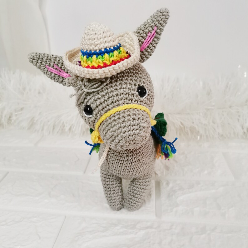 A crocheted grey donkey wearing a small sombrero with a rainbow-coloured brim. The donkey has a yellow bridle and colourful tassels. The ears have pink accents, and the donkey is standing upright.