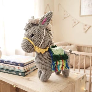 Handmade crochet donkey amigurumi toy with a sombrero hat and colorful saddle standing on a wooden shelf next to children's books.