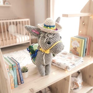 Lifestyle photo of a finished crochet donkey amigurumi toy displayed on a wooden shelf alongside children's books and toys.