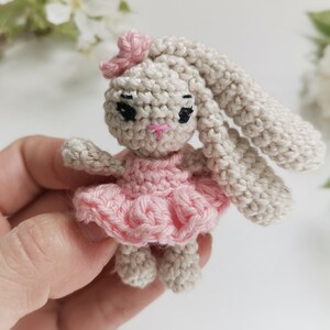 May include: A crocheted bunny doll with long ears, wearing a pink dress and bow. The handmade toy is beige with black eyes and a pink nose. The bunny is a small, soft toy.