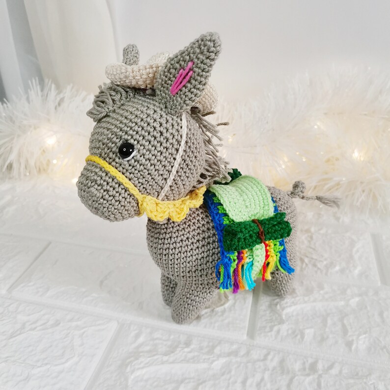 A crocheted grey donkey figurine with a yellow bridle and a green and multicoloured saddle. The donkey has a white mane and tail, and pink accents in its ears. The figurine is handmade and has a whimsical appearance.