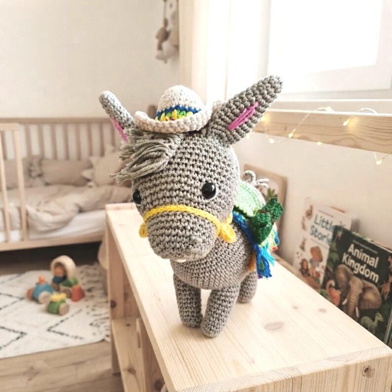 Amigurumi crochet donkey toy styled in a cozy children's bedroom setting with children's books and a wooden house shelf.