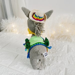 A handmade grey crochet donkey doll wearing a small cream-coloured cowboy hat with a rainbow brim. The donkey also has a green vest, a yellow collar, and a blue and rainbow-coloured shawl. The doll has a white tag that says "Dymas Dolls".