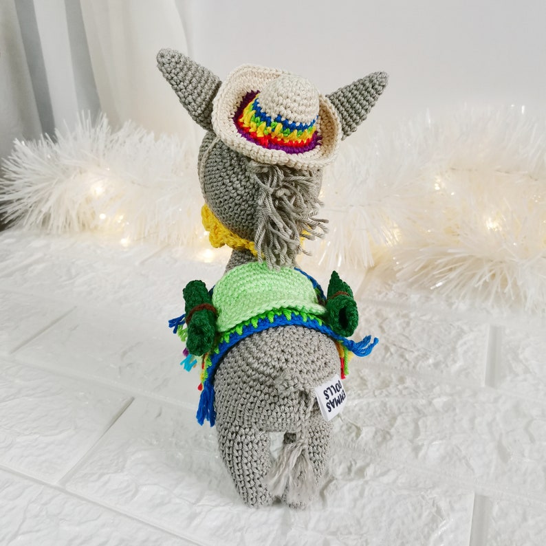 A handmade grey crochet donkey doll wearing a small cream-coloured cowboy hat with a rainbow brim. The donkey also has a green vest, a yellow collar, and a blue and rainbow-coloured shawl. The doll has a white tag that says "Dymas Dolls".