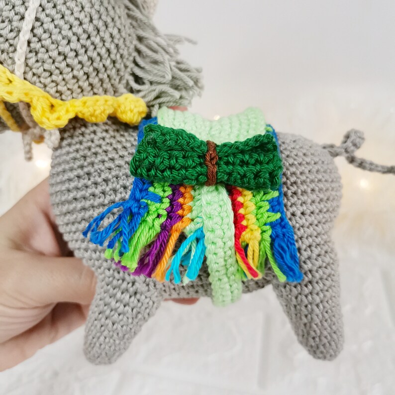 A handmade grey crocheted donkey toy with a colourful fringed blanket and a yellow and white collar. The donkey has grey yarn hair and a green bow on its back. The toy is a soft, plush design.