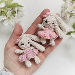 May include: Two small, crocheted bunny dolls. The bunnies are off-white with long ears, black eyes, and pink noses. Each bunny wears a pink dress and a matching bow. The dolls are held in a hand, with white flowers in the background.