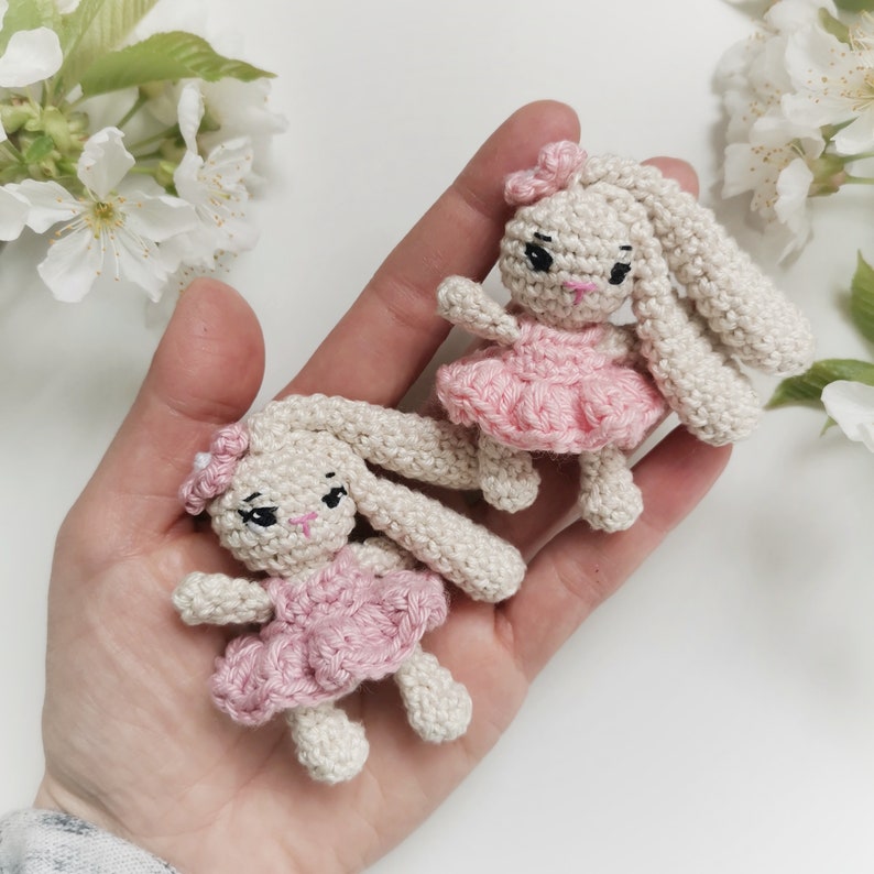 May include: Two small, crocheted bunny dolls. The bunnies are off-white with long ears, black eyes, and pink noses. Each bunny wears a pink dress and a matching bow. The dolls are held in a hand, with white flowers in the background.