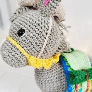A crocheted donkey toy in grey, featuring a yellow bridle and a colourful saddle. The donkey has a white mane and tail, and a black eye. The toy is crafted from soft yarn and is suitable for children.