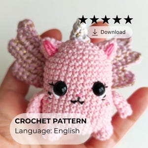 May include: A pink crocheted creature with large black eyes, small pink ears, and gold accents on its wings and horn. The text "CROCHET PATTERN" and "Language: English" are displayed on the image. The creature is held in a hand.