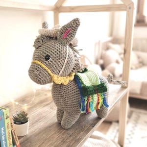 Side profile of a handmade crochet donkey plushie wearing a green saddle with colorful tassels in a nursery room.