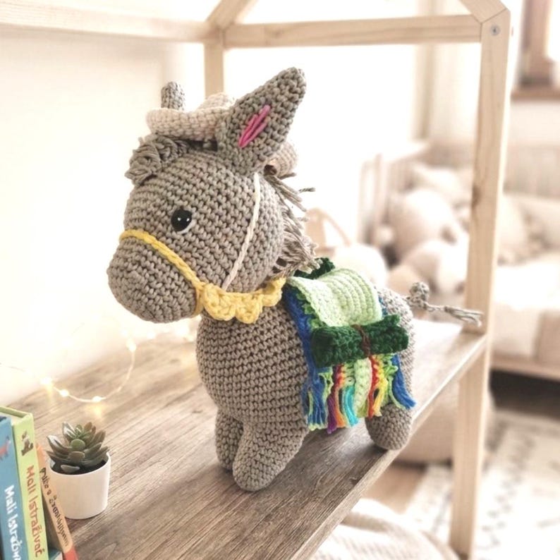 Side profile of a handmade crochet donkey plushie wearing a green saddle with colorful tassels in a nursery room.
