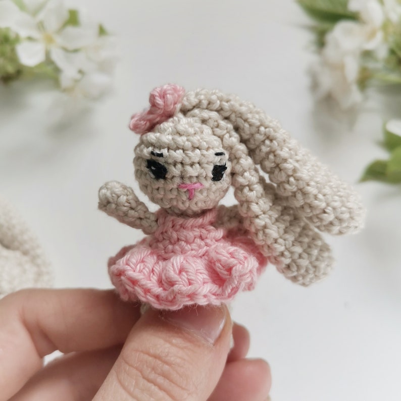 May include: A crocheted bunny doll with long, floppy ears and a pink bow. The bunny wears a pink dress and has black eyes and a pink mouth. The doll is handmade and appears to be a small, decorative item.
