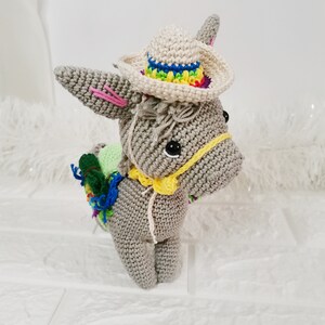 A crocheted grey donkey figurine wearing a cream-coloured sombrero with a rainbow brim. The donkey has a yellow harness and a colourful pack on its back. The donkey is standing on a white surface.