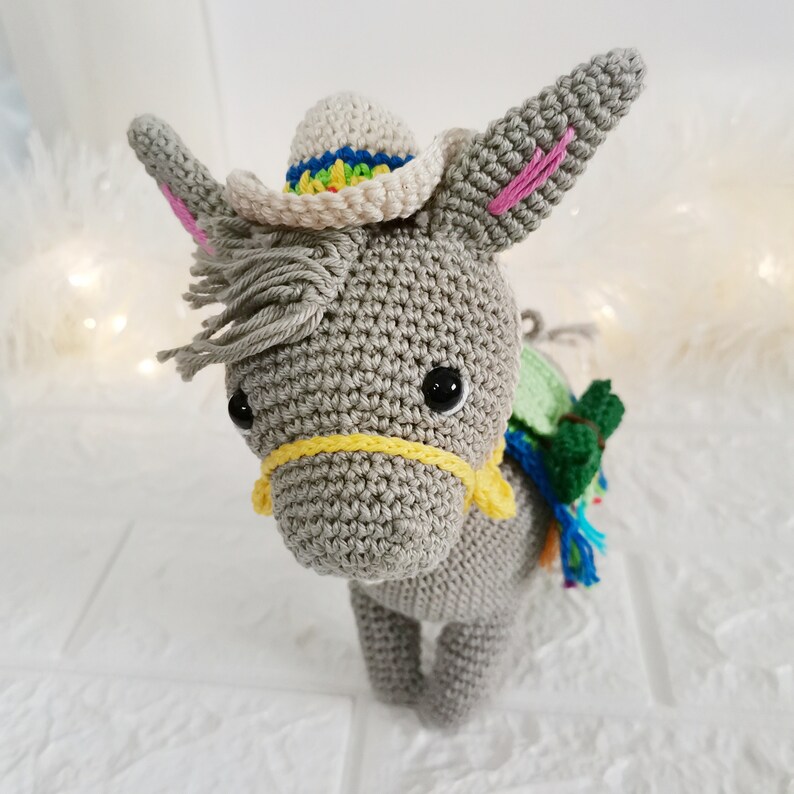 A handmade grey crochet donkey wearing a small white hat with a rainbow brim. The donkey has black eyes, pink inner ears, and a yellow bridle. A green and blue pack is on its back.
